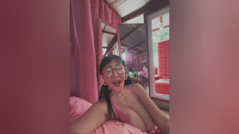 Media: A video of an Asian woman with glasses and long black hair, wearing a pink tank top, lying on a pink bed. Pink curtains and a window with greenery outside create a cozy, intimate bedroom setting.