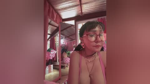 Media: Video of a young Asian woman with a medium complexion, wearing glasses, a pink dress, and multiple necklaces, in a rustic, pink-painted room with wooden walls and a thatched roof, surrounded by floral decorations.