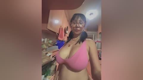 Media: Video of a middle-aged Asian woman with glasses, large breasts, and wearing a pink bra in a cluttered kitchen with beige cabinets, hanging towels, and a white ceiling light.