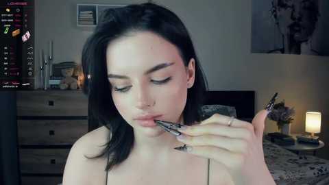 Media: Video of a young woman with pale skin, dark hair, and piercings, applying makeup in a dimly lit bedroom.