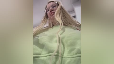 Media: Video of a middle-aged woman with long, blonde hair, wearing large, round glasses and a light green hoodie, standing in a dimly lit room. The image has a soft focus and a slight green tint.