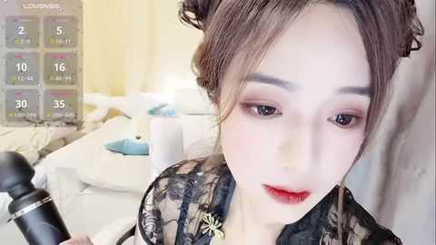 Media: A video of a young East Asian woman with fair skin, styled hair, wearing makeup, and a black floral kimono, sitting in a well-lit, modern room with a white table and blue lamp.