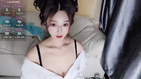 Media: Video of an East Asian woman with fair skin, black hair in a bun, wearing a white shirt and black bra, sitting on a beige couch, surrounded by colorful pillows and a black curtain.