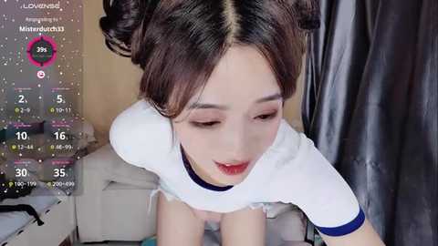 Media: Video of a young Asian woman with dark hair styled in two buns, wearing a white crop top and shorts, kneeling on a bed, with a digital overlay showing her stats.