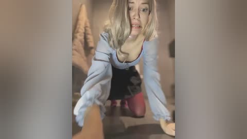 Media: Video of a young woman with blonde hair, wearing a light blue off-shoulder top and black skirt, crawling on a beige carpet, looking up with an open mouth expression.