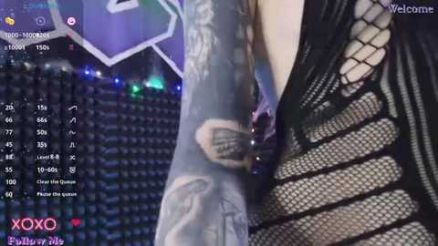 Media: A video of a woman wearing a black fishnet top, showcasing a tattooed arm. The background shows a stadium with a scoreboard and a video overlay of a game, with text \"XOXO\" and \"Welcome\" in the corners.