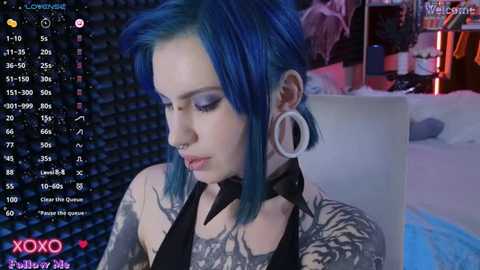 Media: Video of a pale-skinned woman with vibrant blue hair, wearing large white hoop earrings, black choker, and a black top with tattoos. She sits in a dimly-lit room with a bed, shelves, and a window visible in the background.