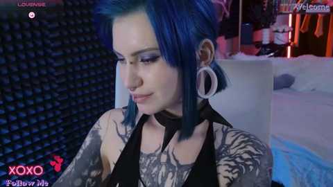 Media: Video of a tattooed woman with blue hair, wearing a black top and choker, in a dimly lit room with a bed and bookshelf in the background.