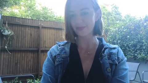 Media: A video of a young woman with shoulder-length brown hair, fair skin, and light makeup, wearing a denim jacket over a black top, standing in a sunlit garden with a wooden fence and greenery background.