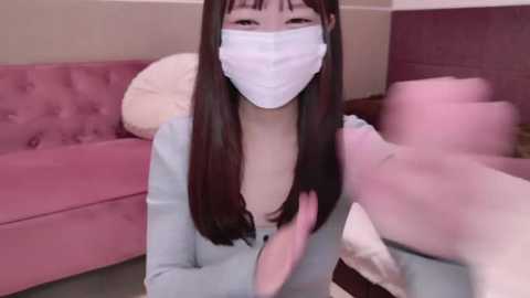 Media: A video of an Asian woman with long black hair, wearing a white mask, light blue cardigan, and a pink shirt. She is indoors, in a room with a pink couch and fluffy pillows.