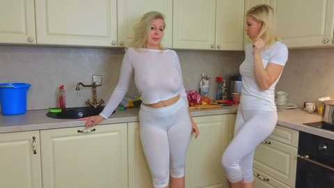 Media: Video of two blonde women in white, semi-transparent crop tops and leggings, standing in a beige kitchen with cabinets, sink, and utensils.