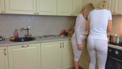 Media: Video of two blonde women in white pajamas standing in a beige kitchen, one washing dishes, the other watching, with cabinets, sink, and countertop visible.