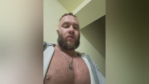 Media: A video of a muscular, bearded, bald white man with a silver chain necklace, wearing an unzipped shirt, standing in a beige-walled room with a corner.