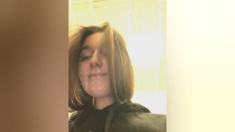 Media: Video of a young woman with light brown hair and a neutral expression, wearing a black cape, with semen on her face, in a poorly lit room with yellowish walls.