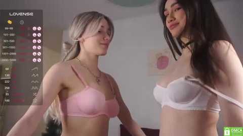 Media: Video of two young women with light skin, one blonde in pink lingerie, the other brunette in white lingerie, smiling in a dimly lit bedroom.