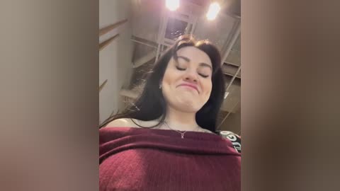 Media: Video of a young woman with long black hair, fair skin, and a round face, wearing a burgundy off-shoulder sweater. She's smiling and tilting her head slightly to the right. The background shows a dimly lit interior with exposed ceiling beams and industrial lighting.