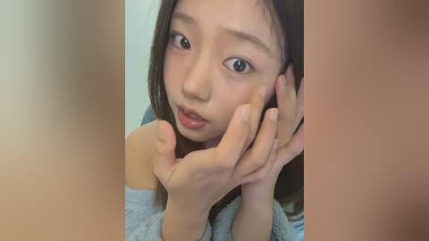 Media: Video of an Asian girl with light skin, long brown hair, and wide, innocent eyes, wearing a gray sweater. She has her hands on her cheeks, looking up with a curious expression. The background is blurred.