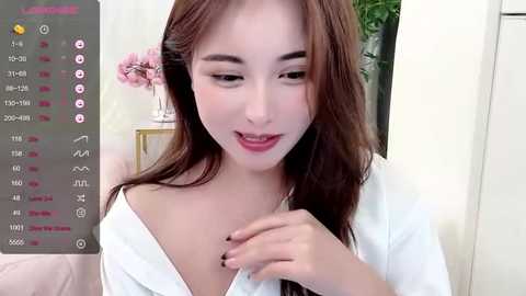 Media: Video of an Asian woman with fair skin and long brown hair, wearing a white robe, smiling, in a room with a pink flower vase and green plants.