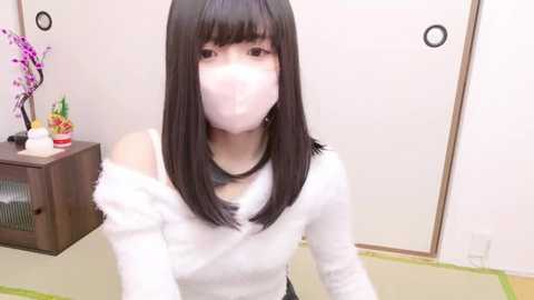 Media: Video of a young Asian woman with straight black hair, wearing a white off-shoulder sweater, face mask, and floral-patterned earrings, sitting on a tatami mat in a traditional Japanese room with a wooden cabinet and floral decorations.