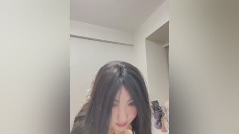 Media: Video of an East Asian woman with long black hair, wearing a dark jacket, standing in a minimalist, bright room with white walls and a closed door. She's slightly out of focus, looking down.