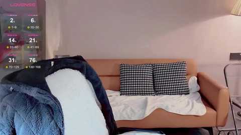 Media: Video of a modern, minimalist bedroom featuring a beige couch with black-and-white checkered pillows, white blanket, and a partially visible person in dark clothing. Background includes a white wall, window, and digital screen displaying a weather forecast.