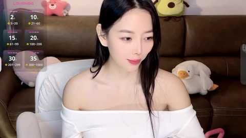 Media: A video of an Asian woman with long black hair, wearing a white off-shoulder dress, sitting on a brown leather couch surrounded by stuffed toys.