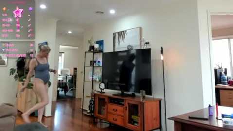 Media: Video of a living room with a woman in a gray one-piece swimsuit dancing near a flat-screen TV, surrounded by a mix of furniture, plants, and electronic devices.