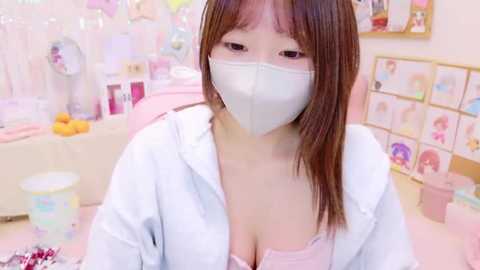 Media: Video of an Asian woman with straight brown hair, wearing a light blue hoodie, pink bra, and white mask, sitting in a pastel-colored room with soft lighting.