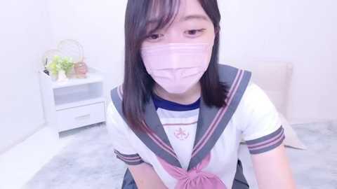 Media: Video of an East Asian woman with light skin and straight black hair, wearing a white schoolgirl uniform, pink surgical mask, and sitting on a light grey carpet in a bright, minimalistic room.