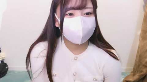 Media: Video of a young East Asian woman with long dark hair, wearing a white face mask and a matching long-sleeved top, seated in a room with beige walls and a brown curtain.