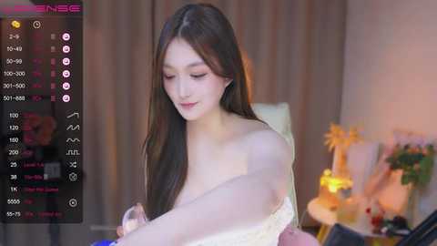 Media: Video of an East Asian woman with long black hair, wearing a light pink top, sitting in a softly lit room with a bed, flowers, and a camera recording her.