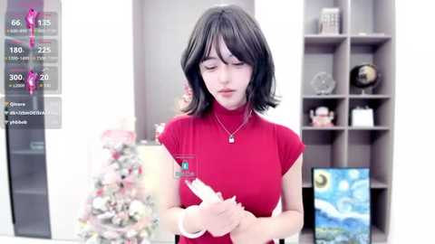 Media: A video of a young Asian woman with short black hair, wearing a red turtleneck, holding a white card. Background features a Christmas tree and a shelf with decorative items.