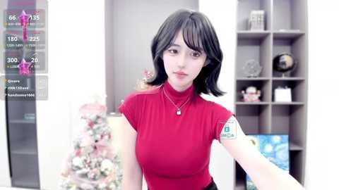 Media: Video of an East Asian woman with short black hair, wearing a red turtleneck, in a modern, minimalist room with gray shelves and a decorated Christmas tree in the background.