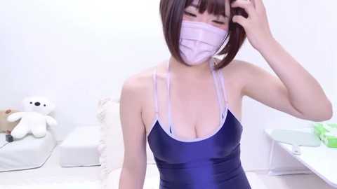 Media: A young East Asian woman with fair skin and short brown hair, wearing a blue one-piece swimsuit with a low-cut neckline and a white face mask, poses in a bright, minimalist room with white furniture and stuffed toys.