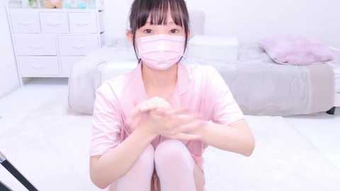 Media: Video of an Asian woman with light skin, sitting on the floor in a bright, minimalist room. She wears a light pink mask, robe, and thigh-high stockings. Background includes a white dresser and a gray couch.