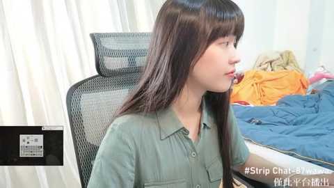 Media: A video shows a young woman with long, straight brown hair, wearing a green button-up shirt, sitting in a black mesh office chair in a messy room with blue and orange bedding.