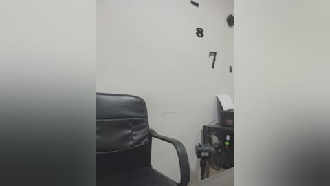 Media: Video of a small, dimly lit interrogation room with a black leather chair and a white wall. Numbers 8 and 7 are written in black on the wall, and a black metal table with a computer monitor and keyboard is visible.