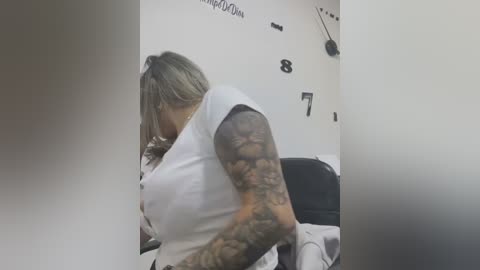 Media: Video of a tattooed woman with short gray hair, wearing a white top, sitting on a chair in a dimly lit room with black wall numbers and a clock.