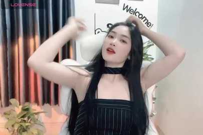 Media: Video of a young East Asian woman with long black hair, wearing a black pinstripe apron, sitting on a white chair, playfully pulling her hair. Background includes a potted plant, curtains, and a \"Welcome\" sign.