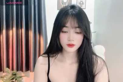 Media: Video of a young East Asian woman with long black hair, fair skin, and red lipstick, wearing a black top, seated indoors with a blurred background featuring a potted plant and curtains.