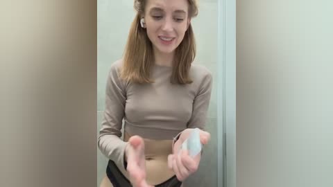 Media: Video of a young, fair-skinned woman with straight, shoulder-length blonde hair, wearing a beige crop top and black panties, standing in a beige-walled bathroom, holding a white cloth.