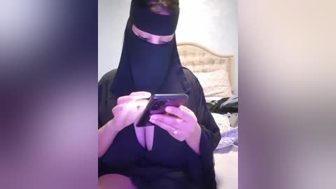 Media: Video of a woman wearing a black burqa with a smartphone, sitting on a bed in a dimly lit bedroom with white walls and a headboard.