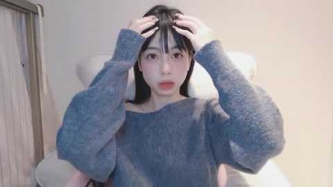 Media: A young Asian woman with long black hair sits on a white chair, wearing a blue, puffy-sleeved sweater. She looks confused, holding her hair with both hands. The background is a plain, light-colored wall with a door visible.