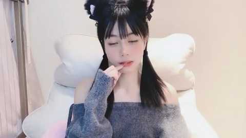 Media: A video of a young Asian woman with long black hair, wearing black cat ears and a grey off-shoulder sweater, sitting on a white chair, looking contemplative, with a hand near her mouth.