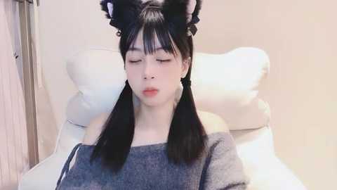 Media: A young Asian woman with pale skin, straight black hair styled in twin pigtails, and cat ears, wearing a gray sweater, lies on a white leather chair in a neutral-toned room.