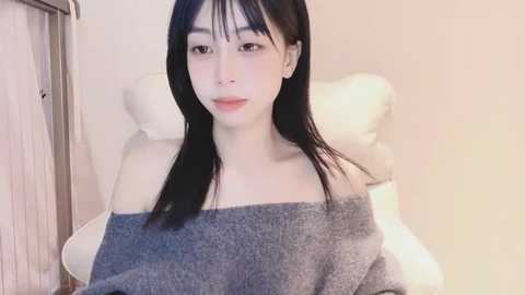 Media: Video of a young Asian woman with long black hair, pale skin, and a slim build, wearing a gray off-shoulder sweater, sitting on a white leather chair in a softly lit room.