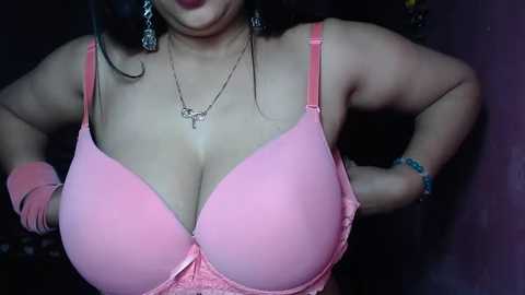 Media: Video of a plus-size woman with medium skin tone, wearing a pink lace bra with visible cleavage, silver necklaces, and bracelets. She stands against a dark background with hands on hips.