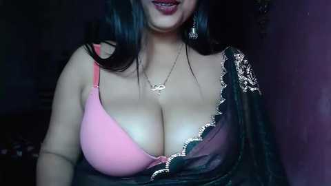 Media: Video of a curvaceous South Asian woman with long black hair, wearing a pink bra with lace detailing and a sheer black blouse with lace trim, revealing ample cleavage.