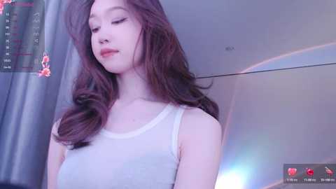 Media: A video of a young East Asian woman with long, wavy brown hair, wearing a white tank top, standing indoors. The background includes a mirror and a partially visible TV screen displaying a live feed.