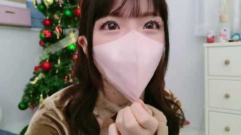 Media: A video of an Asian woman with long dark hair, wearing a pink surgical mask and a brown velvet jacket, standing indoors near a decorated Christmas tree and a white dresser.